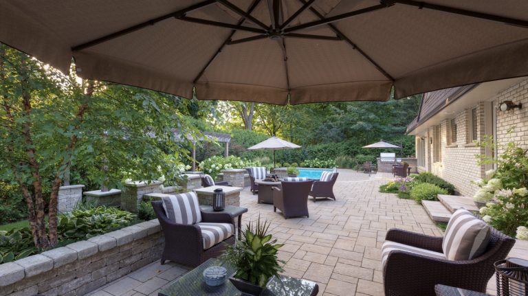 An outdoor living set up with patio furniture, a large umbrella, and a stone patio that leads to an inground pool.