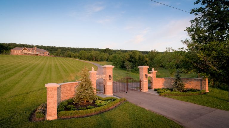 A large stone entry gate that spans over a long driveway leading to a large house.