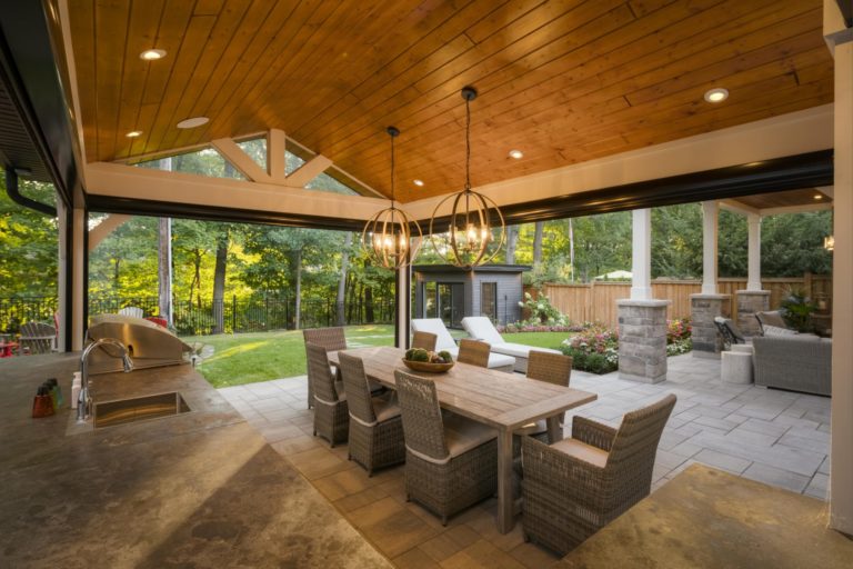 An outdoor kitchen with a dining area and a large overhead covering made of wood with two chandeliers.