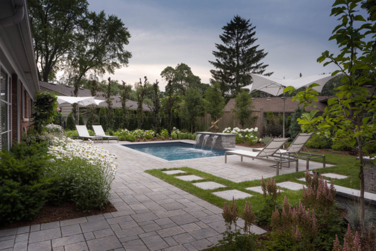 A beautiful inground pool with water features is surrounded with patio stones, beach chairs, and umbrellas.