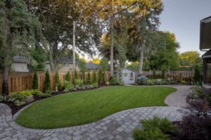 A well-maintained backyard with a small garden shed, lush grass, paved pathways, tall trees, and a wooden fence surrounding the area.