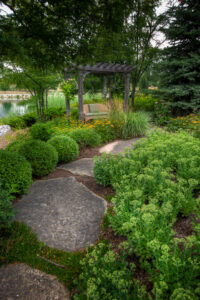 A serene garden scene with blooming flowers and greenery, featuring a wooden pergola and bench near a tranquil pond under trees.