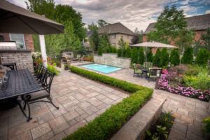 A well-maintained backyard features a small swimming pool, patio dining area, colorful flowers, and lush greenery under a partly cloudy sky.