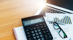 A calculator displaying "2025" rests on a desk with a laptop, eyeglasses, and financial charts, suggesting an analysis or planning session.